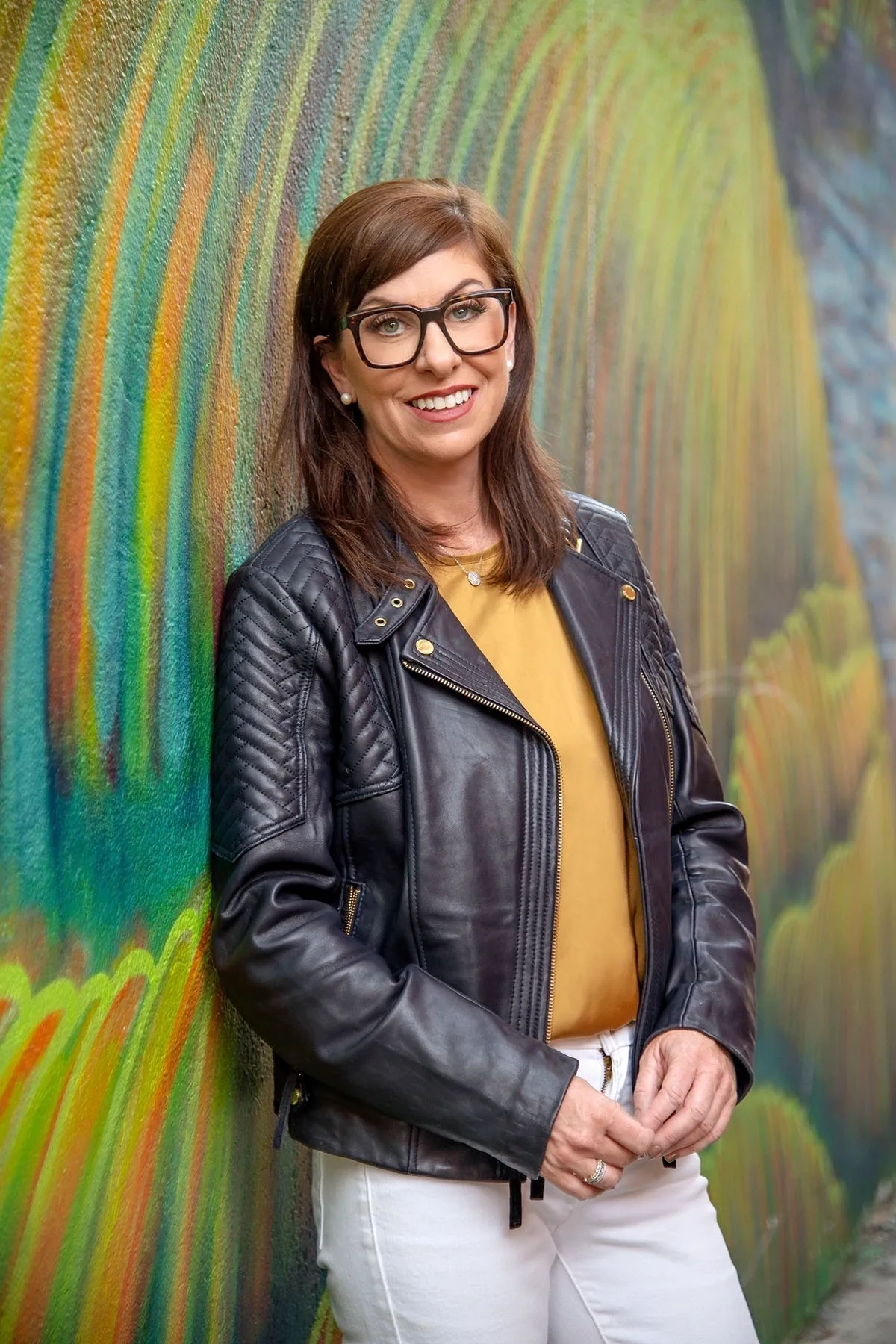 A woman in glasses and leather jacket standing next to a wall.